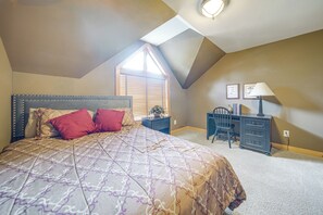 Um quarto com teto inclinado, uma cama com roupa de cama estampada, uma escrivaninha com luminária e uma janela com persianas.