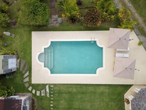 Eine Vogelperspektive auf einen rechteckigen Pool mit einer umlaufenden Liegefläche, umgeben von gepflegten Rasenflächen und verschiedenen Pflanzen.