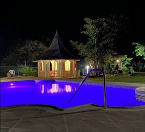 Een zwembad met een blauw verlichte rand, een kleine, gazebo-achtige constructie met een puntig dak, en een metalen trap die naar het zwembad leidt.