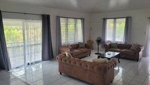Ein geräumiges Wohnzimmer mit zwei braunen Sofas, einem Couchtisch und einem Standventilator. Große Fenster mit Vorhängen sorgen für natürliches Licht und Blick auf die grüne Umgebung.