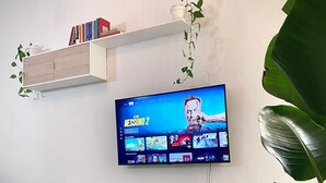Ein flachbildfernseher an der Wand, darunter ein Holzkabinett und rechts eine Topfpflanze.