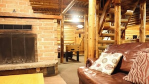 Interior de una cabaña de troncos con chimenea de ladrillo, sofá de cuero y vigas de madera.