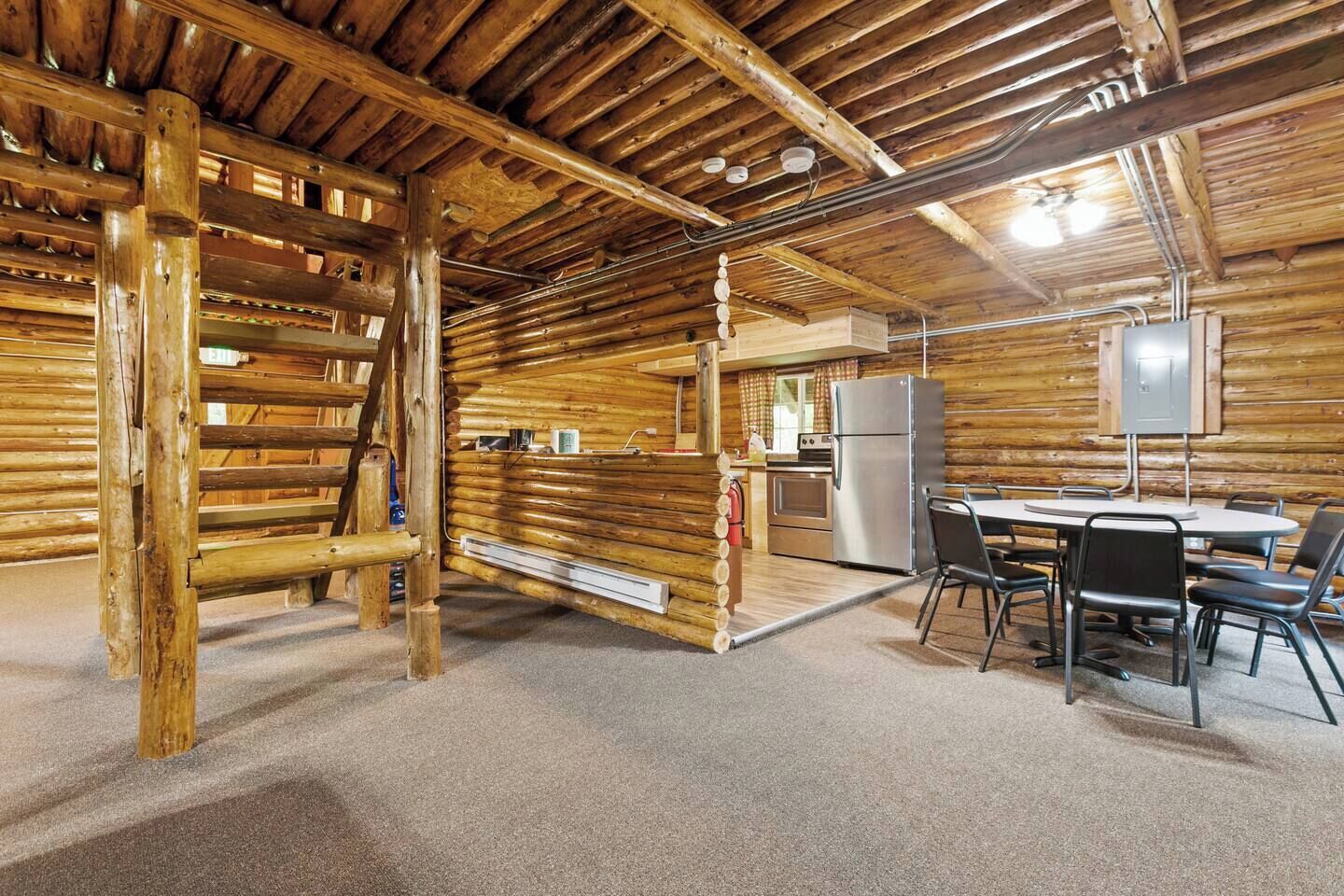 A log cabin interior with a dining area, kitchen, and a wooden ladder.