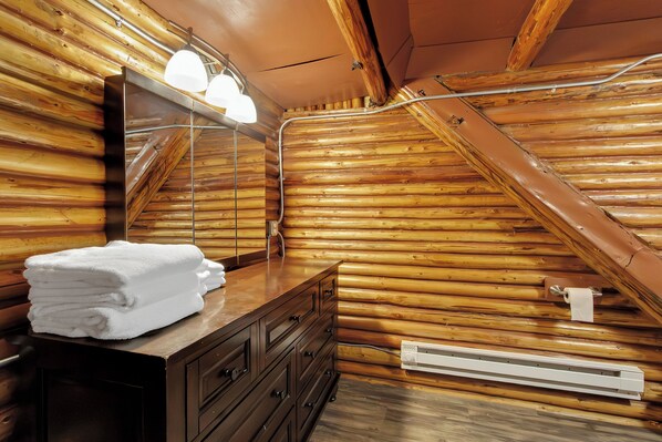 A wooden cabin bathroom with a wooden sloped ceiling, a vanity with a mirror, a towel stack, and a towel dispenser.