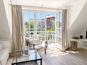 Ein Zimmer mit Glastisch, einer Couch, einem Sessel, einem Fernseher und einem Balkon mit Blick auf Häuser und Grünflächen.