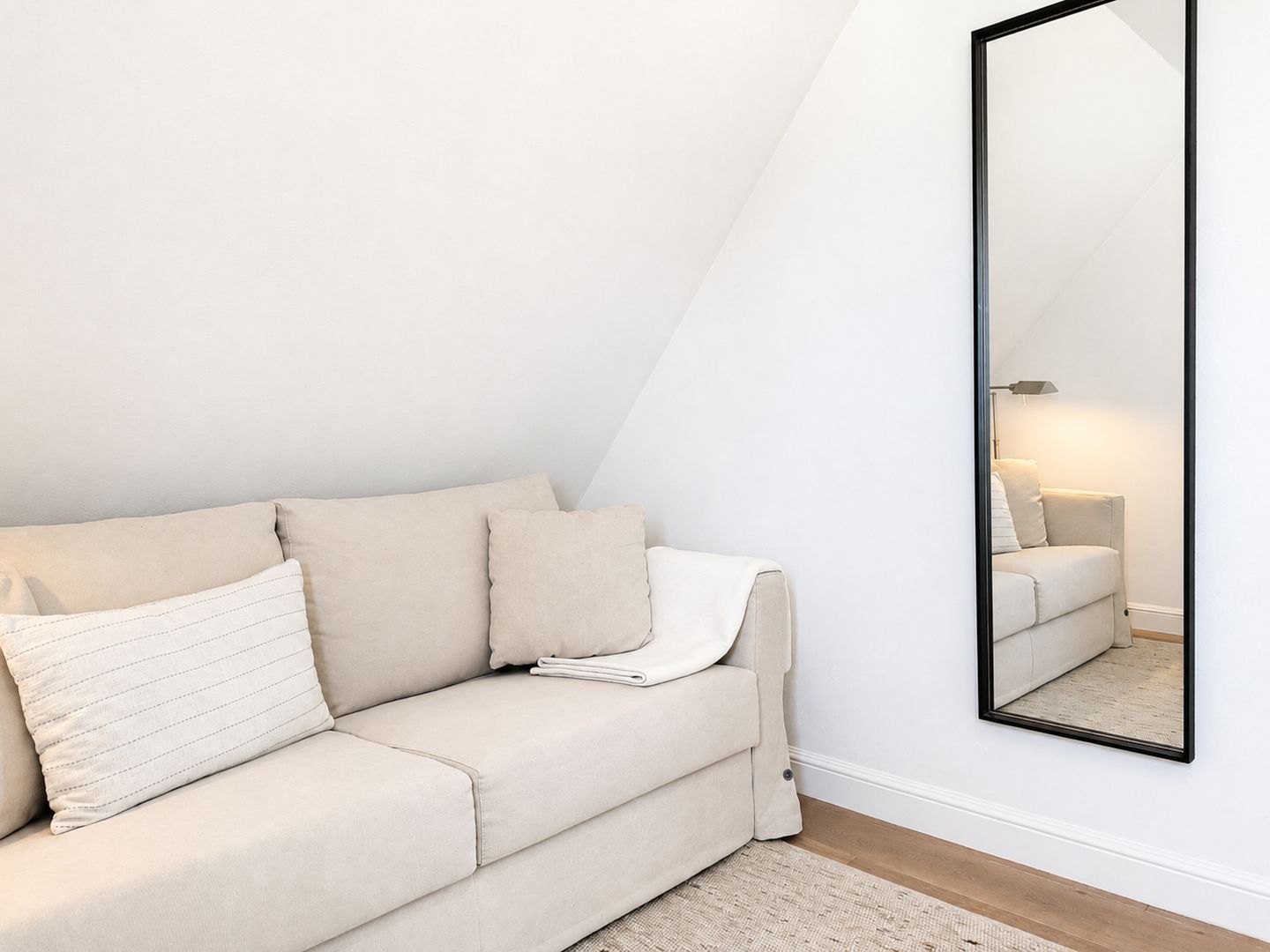 A modern, minimalist living room with a beige sofa, a large mirror, and a simple floor lamp.