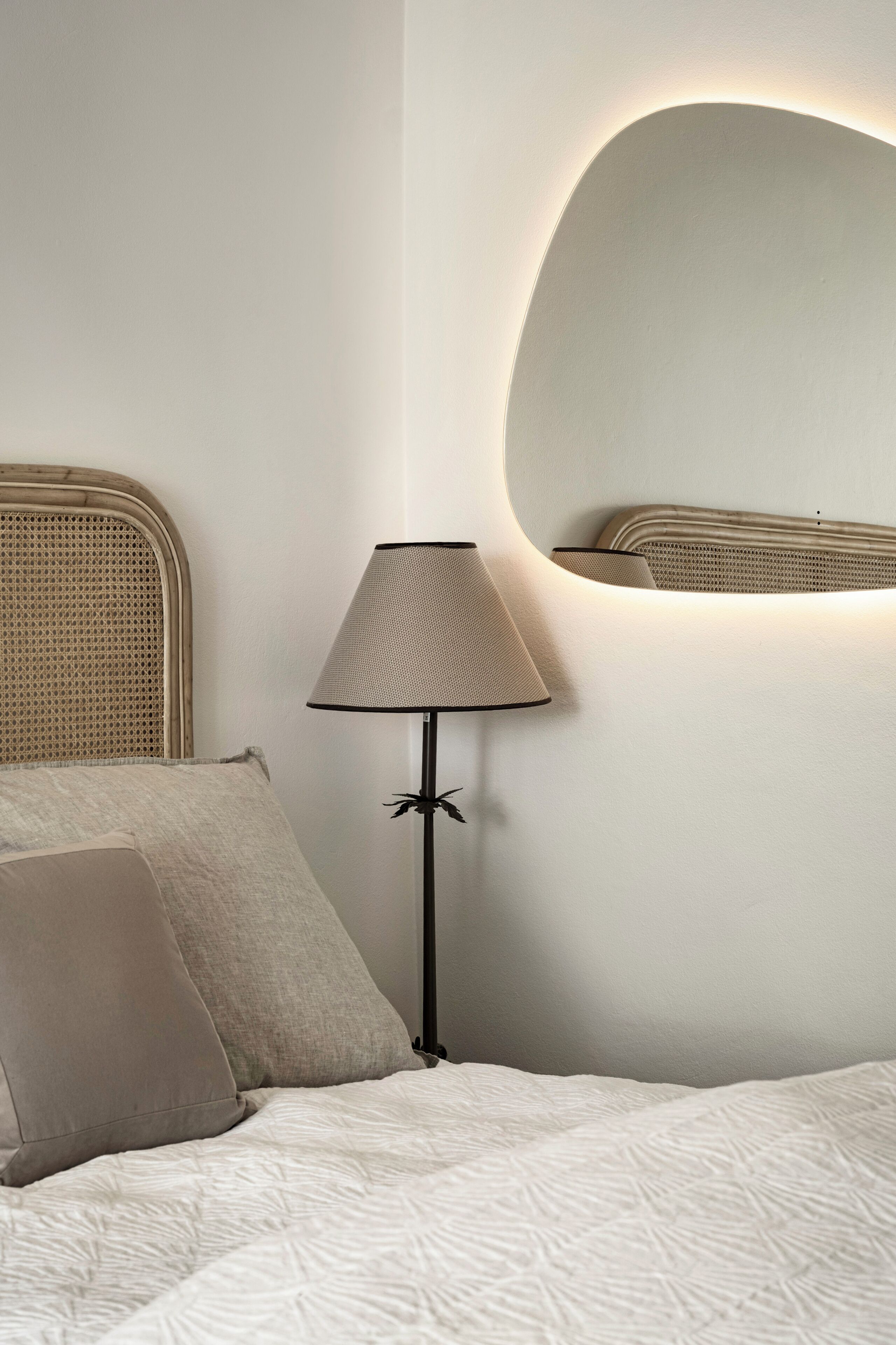 Een bed met een ribbelige witte dekbedovertrek, een lamp met een gevlochten kap en een ronde spiegel aan de muur.