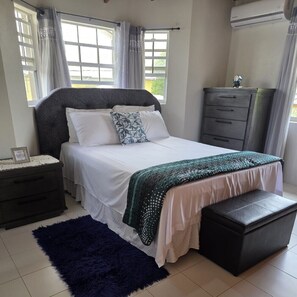 A bedroom with a bed, a nightstand, a dresser, a bench, and a window with curtains. - Spacious 2BR Home with Private Parking & Large Patio. (Frere Pilgrim)