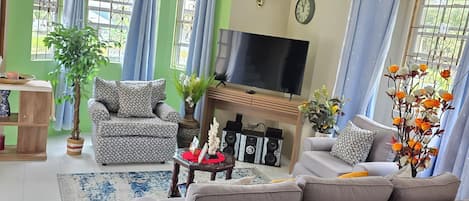 A living room with a grey sofa, a TV, a ceiling fan, and a large window with curtains.