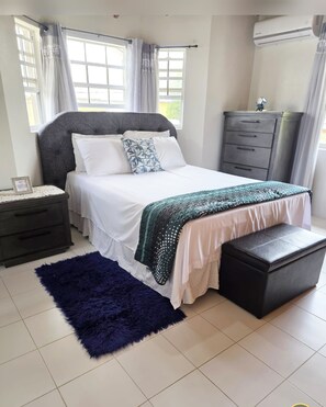 A bedroom with a bed, a nightstand, a dresser, a bench, and an air conditioning unit. - Escape the Cold: Shavaz Paradize 2BR Home Near Golf & Beach, Free Parking & WiFi (Frere Pilgrim)