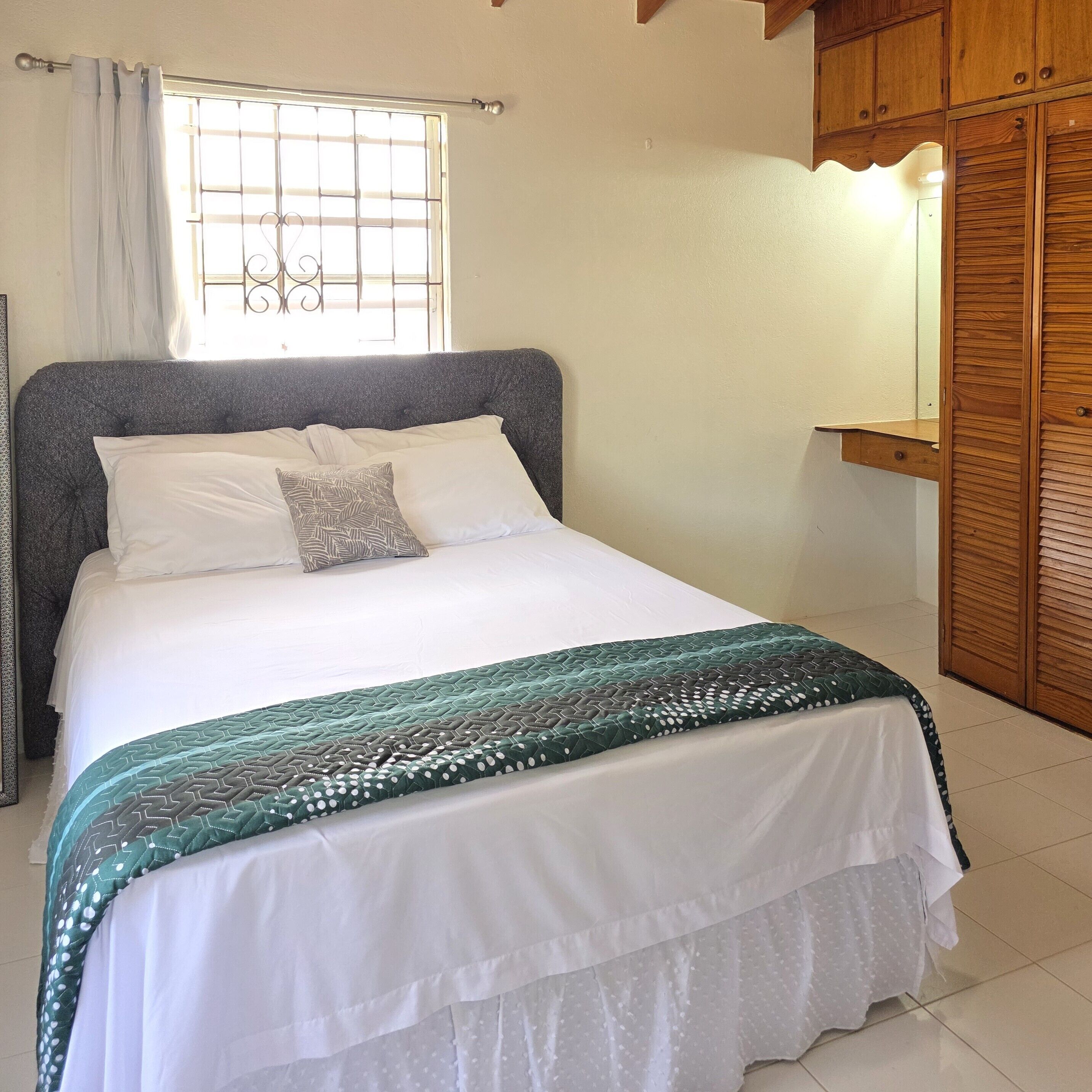 Een netjes opgemaakt bed met een bedsprei met patroon, een raam met tralies en een houten garderobe.