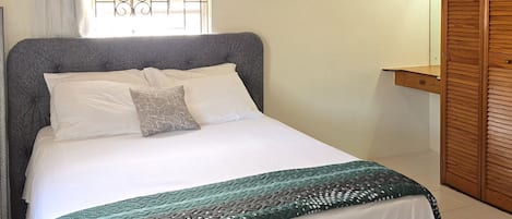A neatly made bed with a patterned bedspread, a window with bars, and a wooden wardrobe.