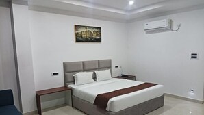 A modern hotel room with a large bed, bedside tables, a nightstand, a wall-mounted air conditioner, and a painting on the wall. - Hotel RKD Inn (Guwahati)