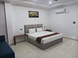 A modern hotel room with a large bed, bedside tables, a nightstand, a wall-mounted air conditioner, and a painting on the wall. - Hotel RKD Inn (Guwahati)