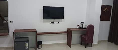 Una habitación con televisión de pantalla plana montada en la pared, un escritorio con una silla, un refrigerador pequeño y un cesto de basura.