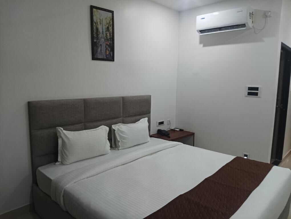 A hotel room with a bed, a nightstand, an air conditioning unit, and a painting on the wall.