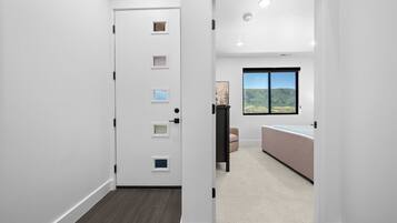 A hallway leading to a bedroom with a large window, a door with multiple peepholes, and a carpeted floor.
