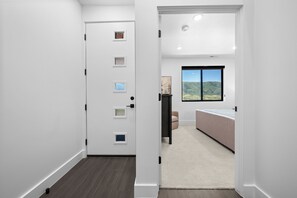 Un pasillo que conduce a un dormitorio con un ventanal grande, una puerta con varios mirillas y piso alfombrado.