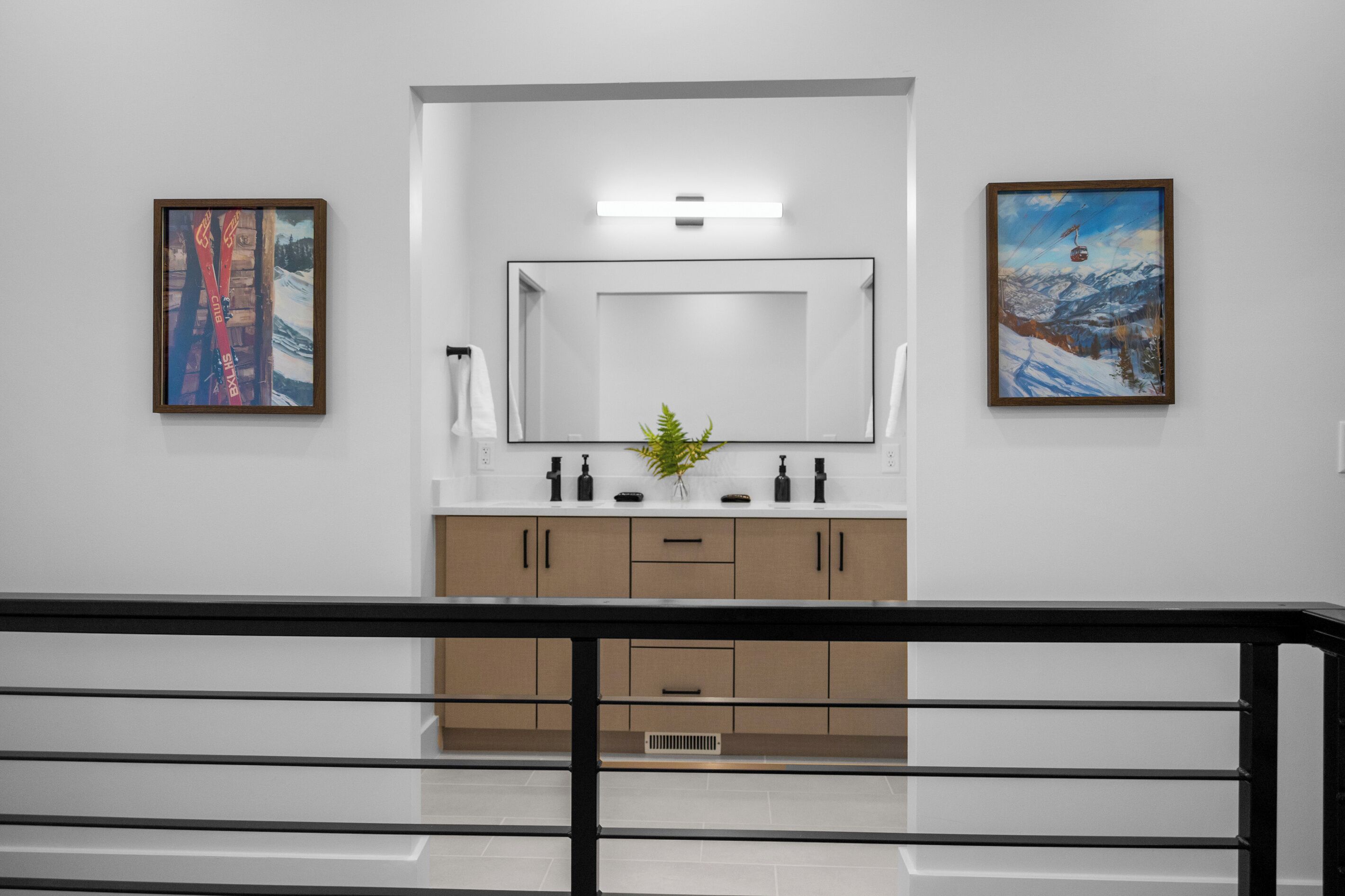 A bathroom with a large mirror, two framed artworks, a sink with toiletries, and a plant.