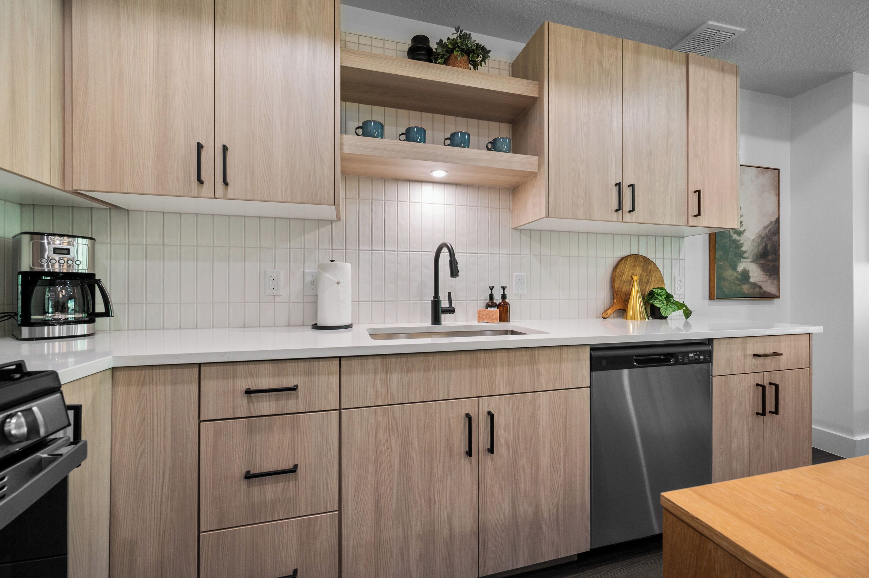 A modern kitchen with light wood cabinets, a stainless steel dishwasher, and a coffee maker.