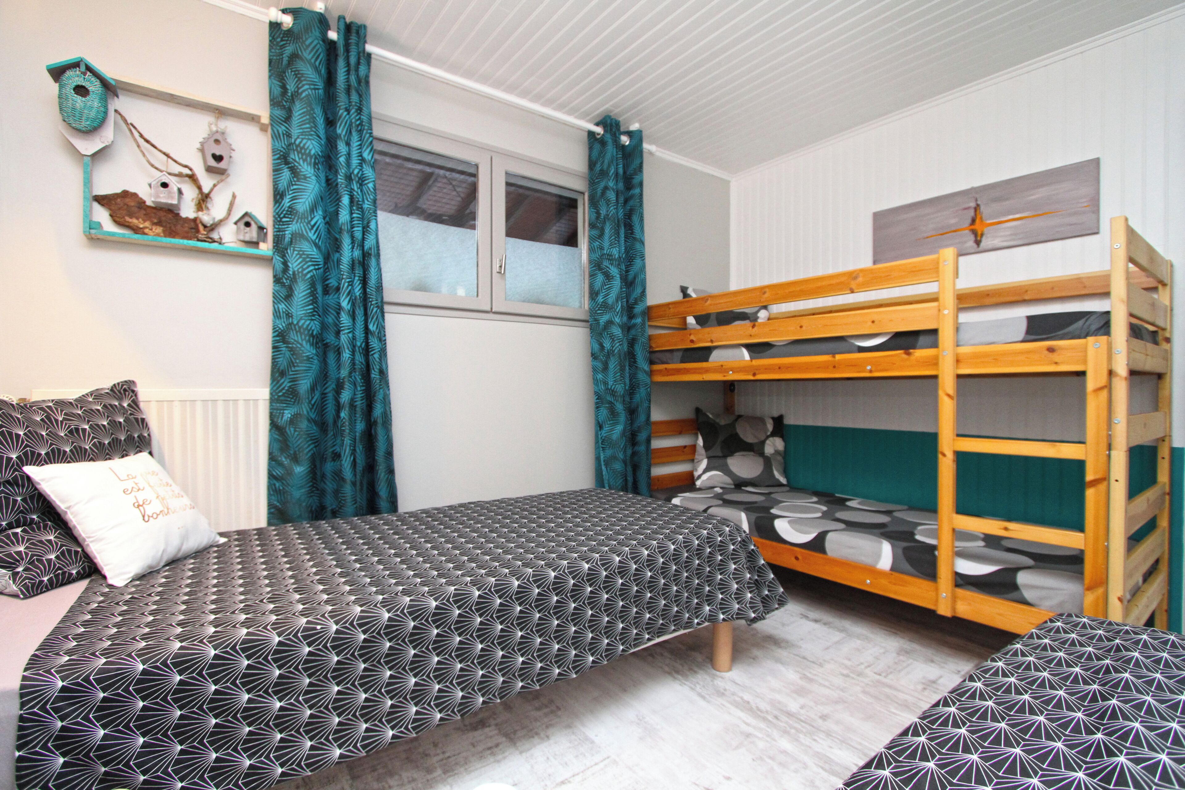 A bedroom with a bunk bed, a single bed, patterned bedding, a window with curtains, and wall decorations including birdhouses.