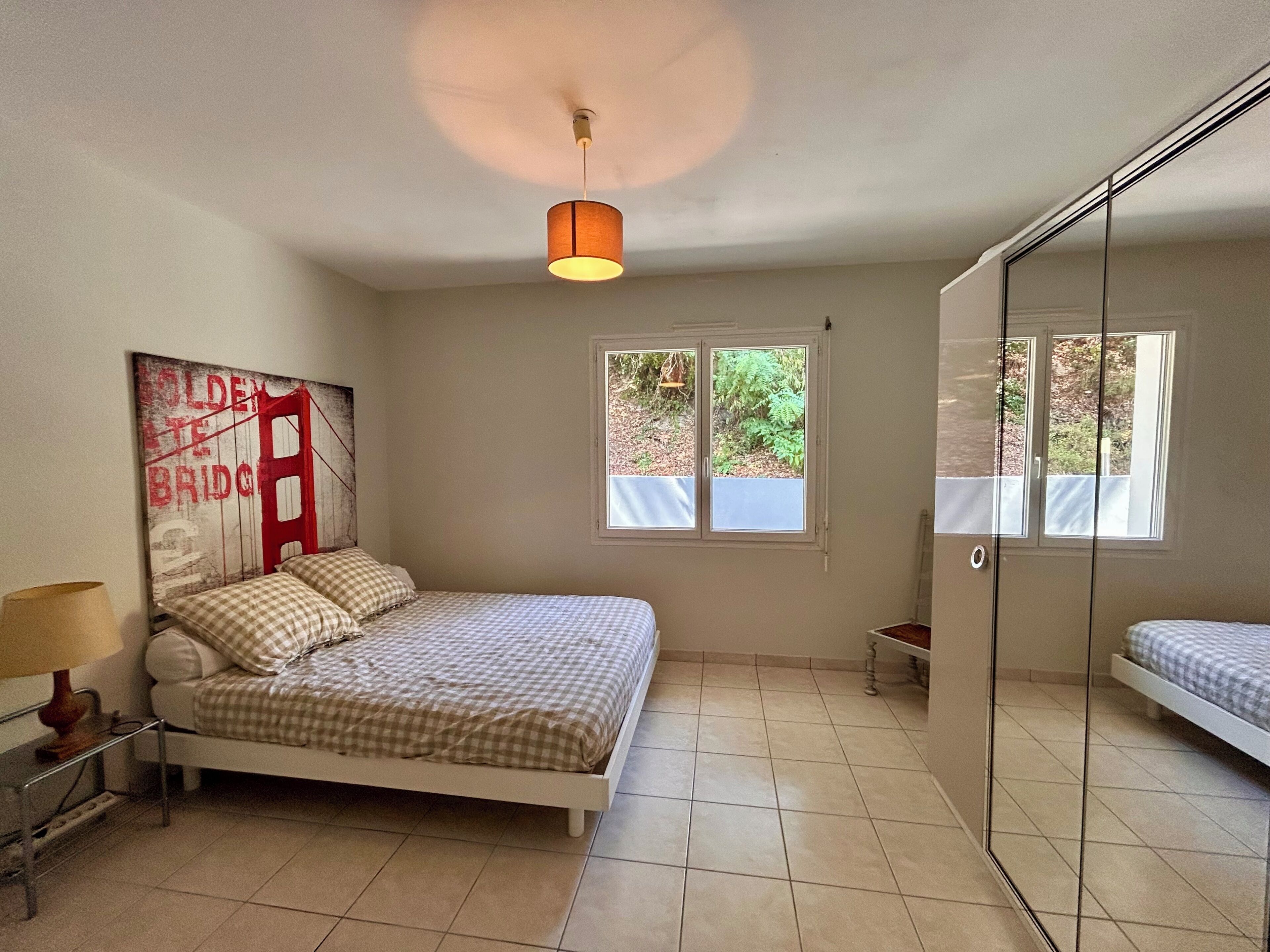 Une chambre à coucher avec un lit, une table de chevet, une lampe, une fenêtre et une porte dotée d’un miroir.