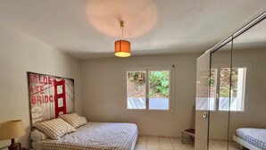 Ein Schlafzimmer mit einem Bett, einem Nachttisch, einer Lampe, einem Fenster und einer Tür mit einem Spiegel.