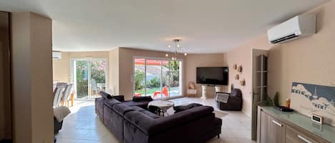 Un salon moderne avec un canapé d’angle, un téléviseur à écran plat et un coin repas avec des chaises.
