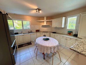 A kitchen with white cabinets, a dining table with a floral tablecloth, and a window with a view of greenery. - Villa with a pool in the heart of Pyla-sur-Mer / La Teste-de-Buch / Arcachon (La Teste-de-Buch)