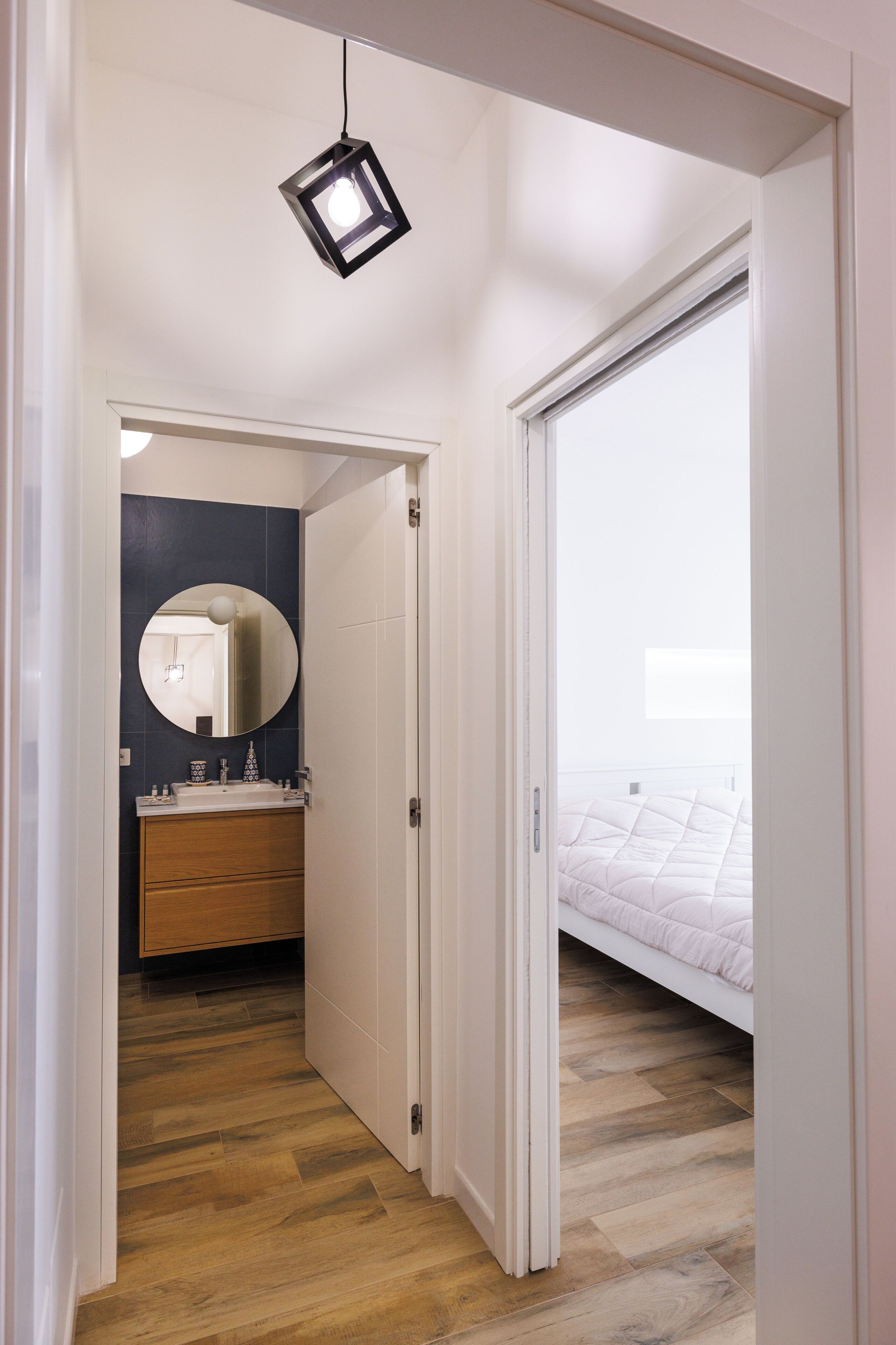A hallway leading to a bedroom with a bed, a bathroom with a mirror, and a hanging light fixture.