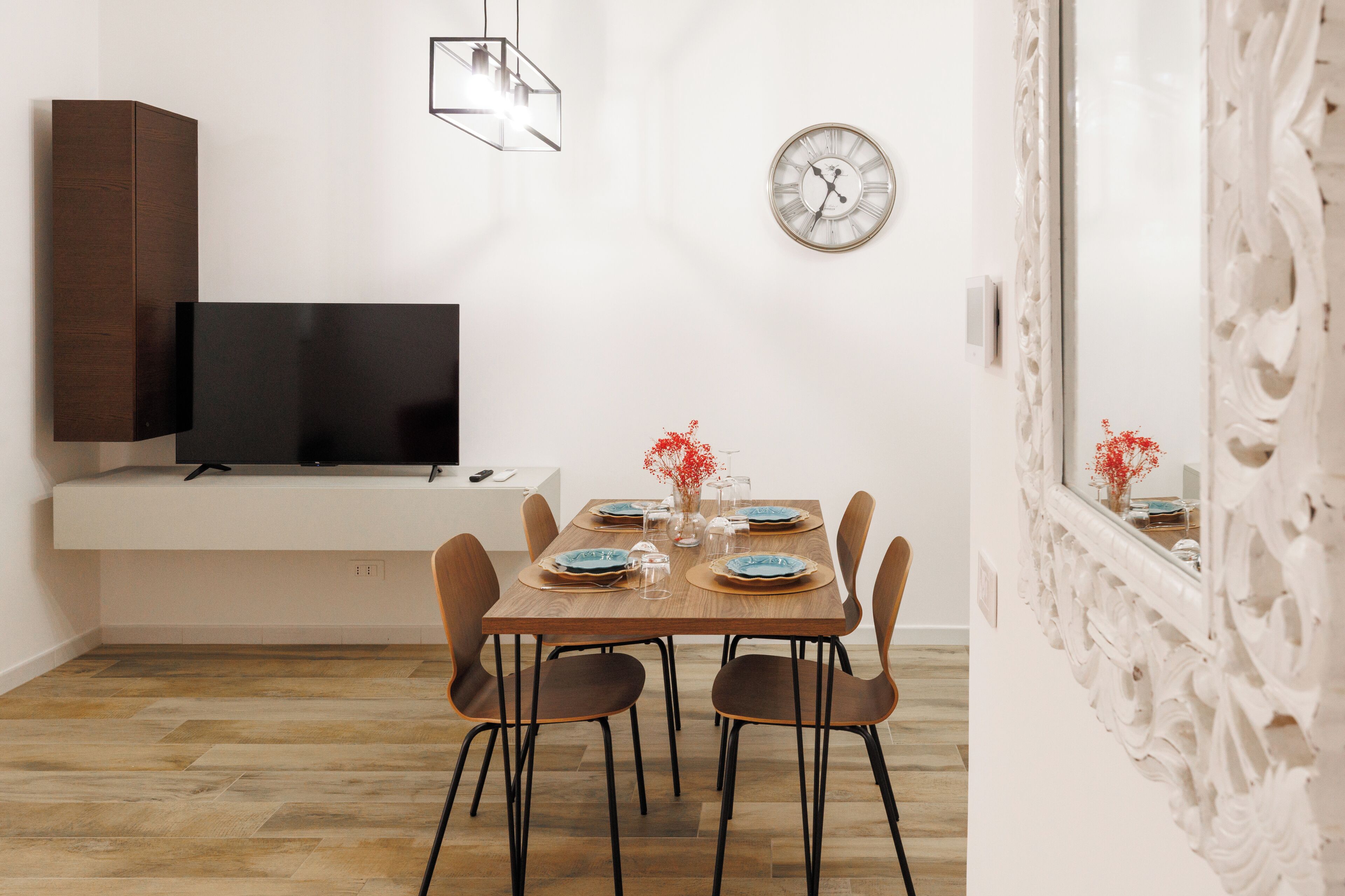 A modern dining area with a wooden table set for four, a flat-screen TV on a stand, a wall clock, and a decorative mirror.