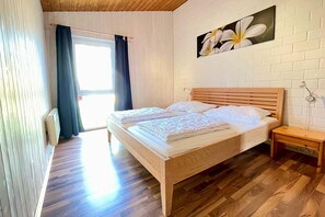 A bedroom with a wooden bed, a nightstand, a wall-mounted lamp, and a window with curtains. - Moderner Doppelbungalow mit Terrasse und Grill im Ferienpark Mirow (Granzow)