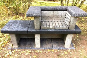 A stone bench with a metal grill structure on top, set in a park with fallen leaves. - Moderner Doppelbungalow mit Terrasse und Grill im Ferienpark Mirow (Granzow)