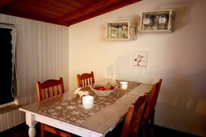 Un coin repas avec une table dressée pour quatre personnes, entourée de chaises en bois. On y trouve deux boîtiers électriques fixés au mur et un tableau encadré.