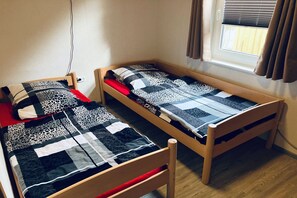 Ein Einzelbett aus Holz mit einem roten Kopfkissen und einer gemusterten Decke, eine Steckdose an der Wand und ein Fenster mit Vorhängen.