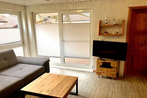 A living room with a sofa, a wooden coffee table, a television on a wooden stand, and a sliding glass door leading to an outdoor area. - Urlaub am Wasser - Doppelbungalow mit Terrasse im Ferienpark Mirow (Granzow)