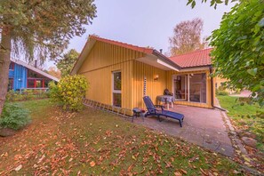 A yellow wooden house with a red roof, a patio with two lounge chairs, and a small table. - Skandinavischer Doppelbungalow mit Terrasse im Ferienpark Mirow (Granzow)
