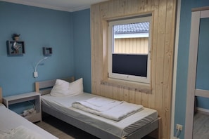 Ein Einzelbett mit Holzvorhang, ein Nachttisch, eine kleine Ablagefläche mit Dekoration und ein Fenster mit Jalousien.