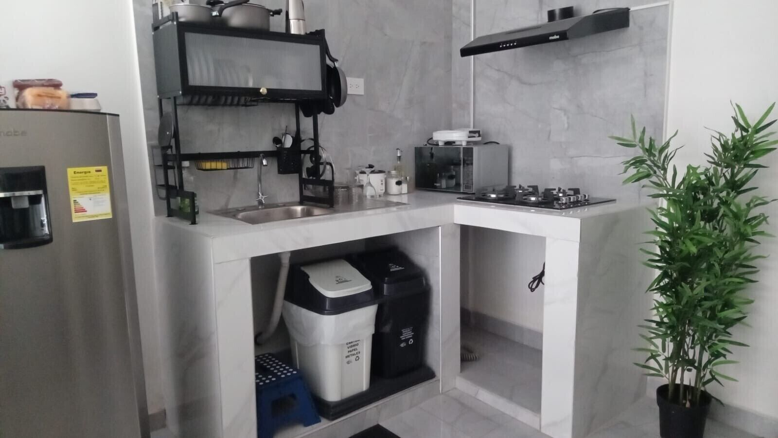 A compact kitchen with a built-in microwave, stove, and sink. There is a trash bin and a small shelf with kitchen items.