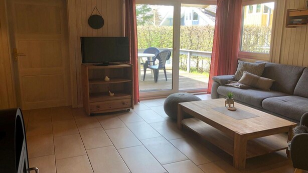 A living room with a sofa, a coffee table, a television on a stand, and a sliding glass door leading to an outdoor area with a patio table and chairs. - Double bungalow in country house style with terrace in the Mirow holiday park (Granzow)