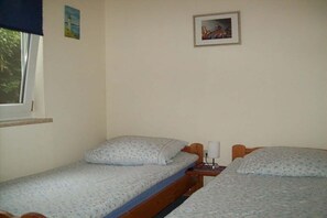 Ein Zimmer mit zwei Einzelbetten, einem Fenster, einem Bild an der Wand und einem kleinen Tisch mit einer Lampe.