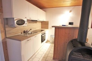 A compact kitchen with white cabinets, a microwave, a sink, an oven, and a wood-burning stove. - Schöner Doppelbungalow mit Terrasse im Ferienpark Mirow (Granzow)