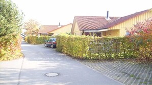 Uma rua residencial com casas, um carro estacionado na lateral e uma tampa de bueiro na via.