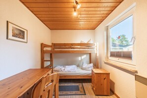 Ein Schlafraum mit Etagenbetten, einem Holztisch und Stuhl, einem gerahmten Bild an der Wand und einem Fenster mit Blick auf Bäume.