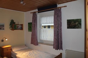 A bedroom with a single bed, a nightstand, a wall-mounted lamp, a window with curtains, and two framed pictures. - Praktischer Doppelbungalow mit Terrasse im Ferienpark Mirow (Granzow)