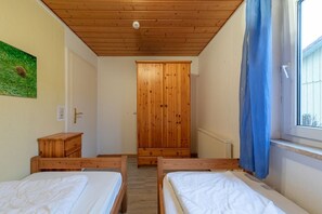 Ein Zimmer mit zwei Holzbetten, einem Holzschrank, einem Heizkörper und einem Fenster mit blauer Gardine.