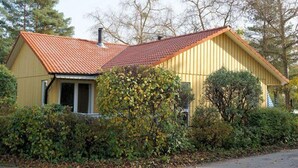 Ein gelbes Holzhaus mit rotem Ziegeldach, umgeben von dichtem Buschwerk und Bäumen.