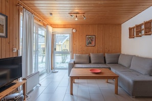 Ein modernes Wohnzimmer mit Holzboden, einem großen Sofa, einem Couchtisch, einem Fernseher und einer Glastür zum Garten.