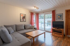 A living room with a grey sofa, a wooden coffee table, a television on a wooden stand, and a sliding glass door leading outside. - Gemütlich eingerichteter Doppelbungalow mit Garten in Mirow (Granzow)
