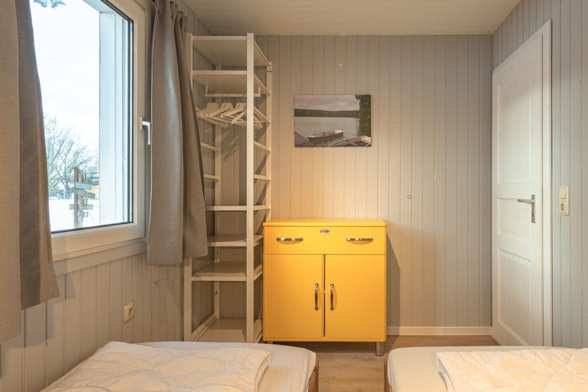 A room with two beds, a yellow wardrobe, a window with curtains, and a painting on the wall.